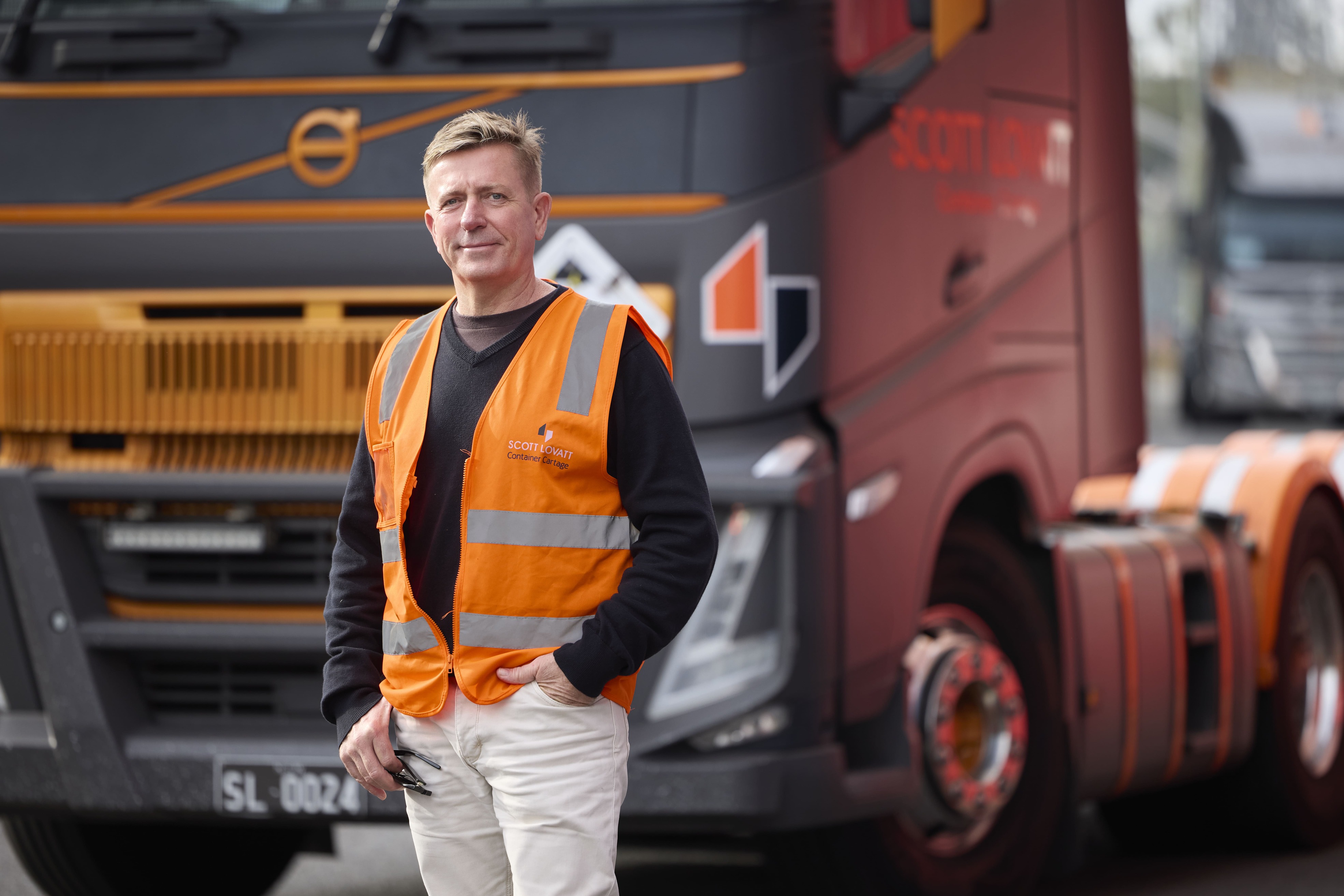 Man in orange safety vest standing in front of a large truck with company logos.