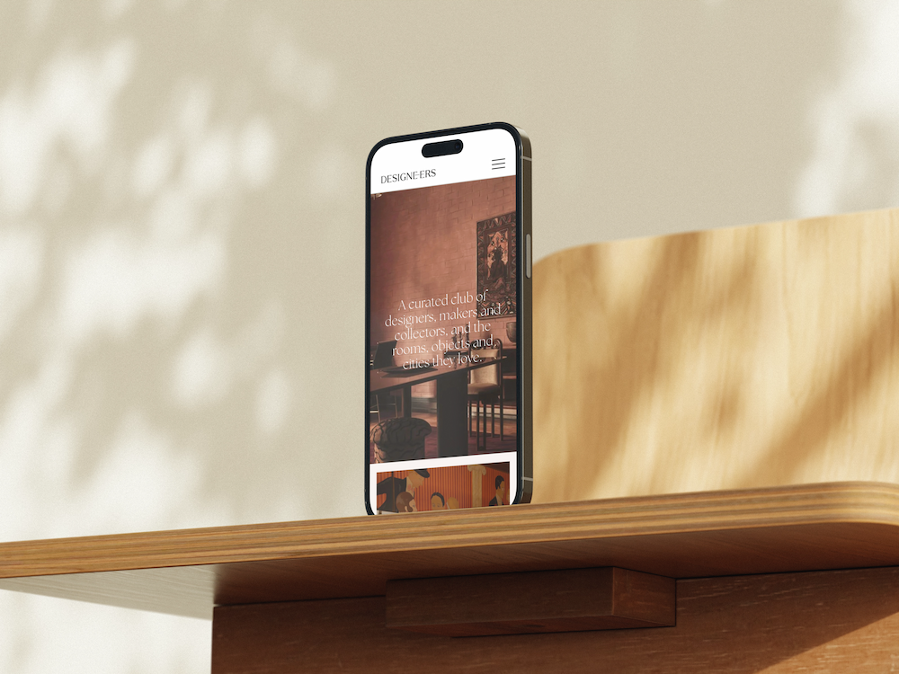 Designeers Club's homepage on a phone against a wooden surface, "A curated club of designers, makers"