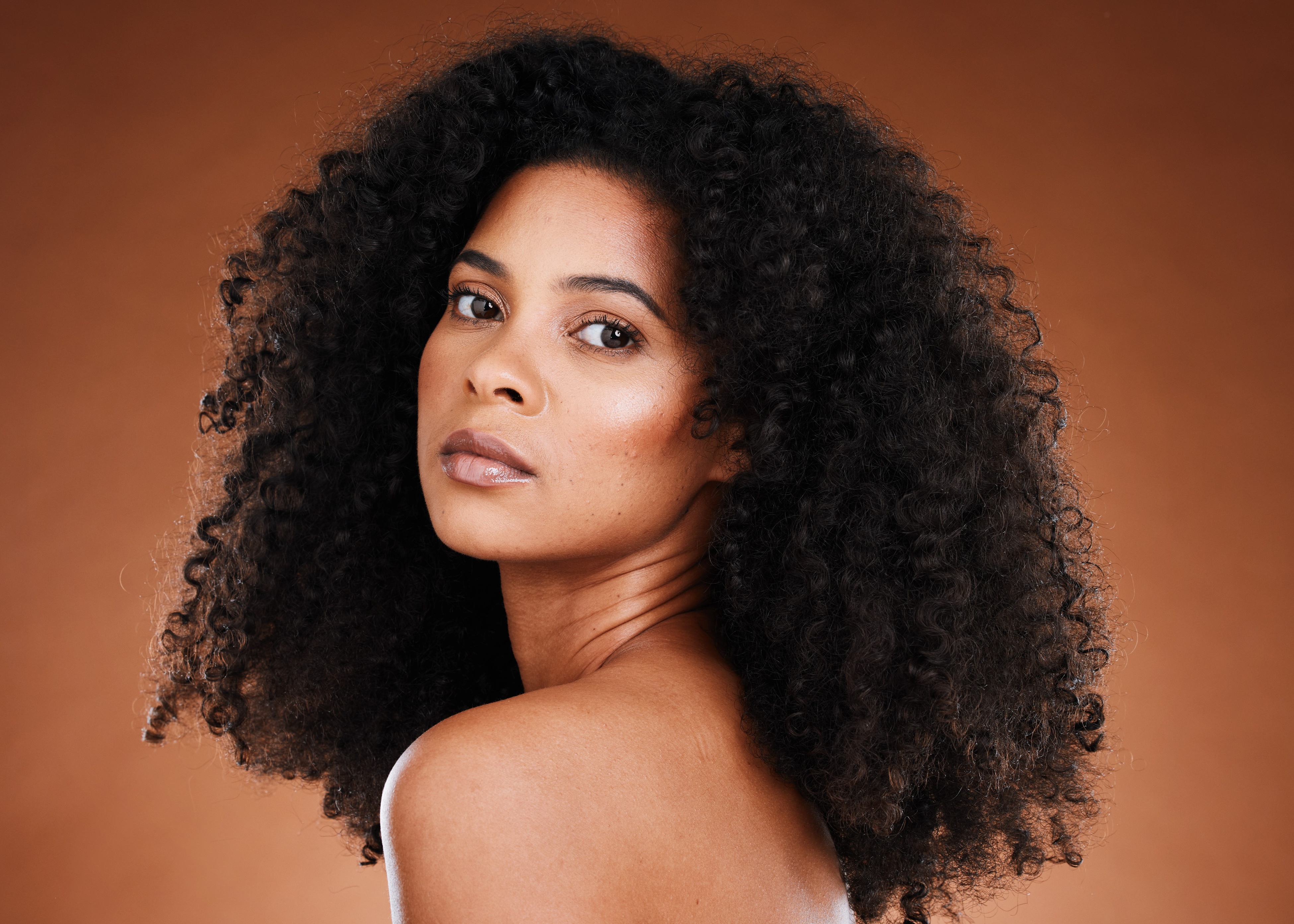 Portrait of a woman with natural curly hair looking over her shoulder against a warm brown background.