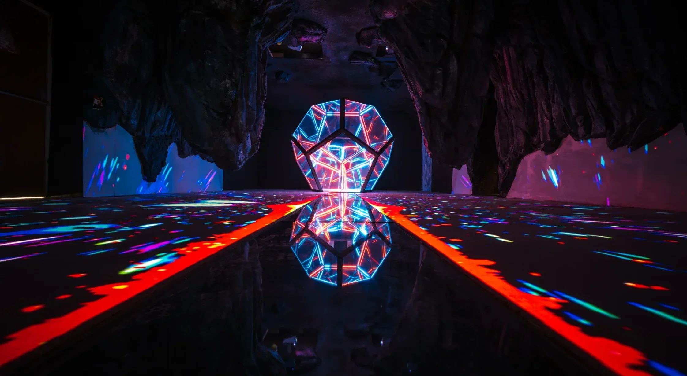 Neon-lit geometric sculpture reflecting on a glossy black floor inside a dark cave.