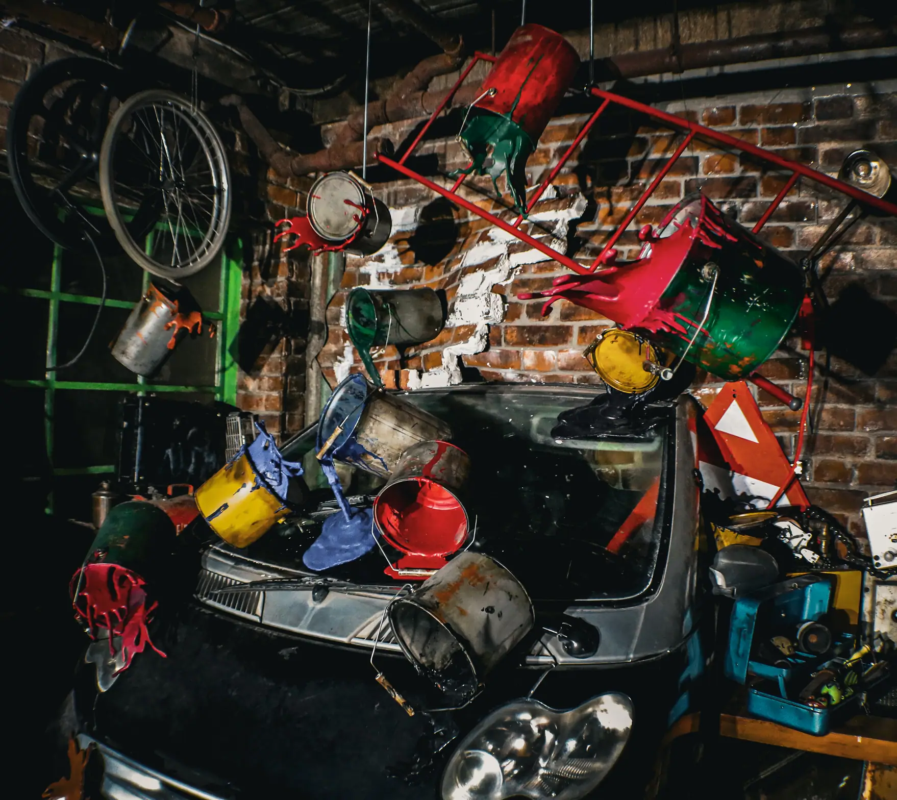 Car in a garage with paint cans spilling various colors suspended around it and bicycle wheels on a brick wall.