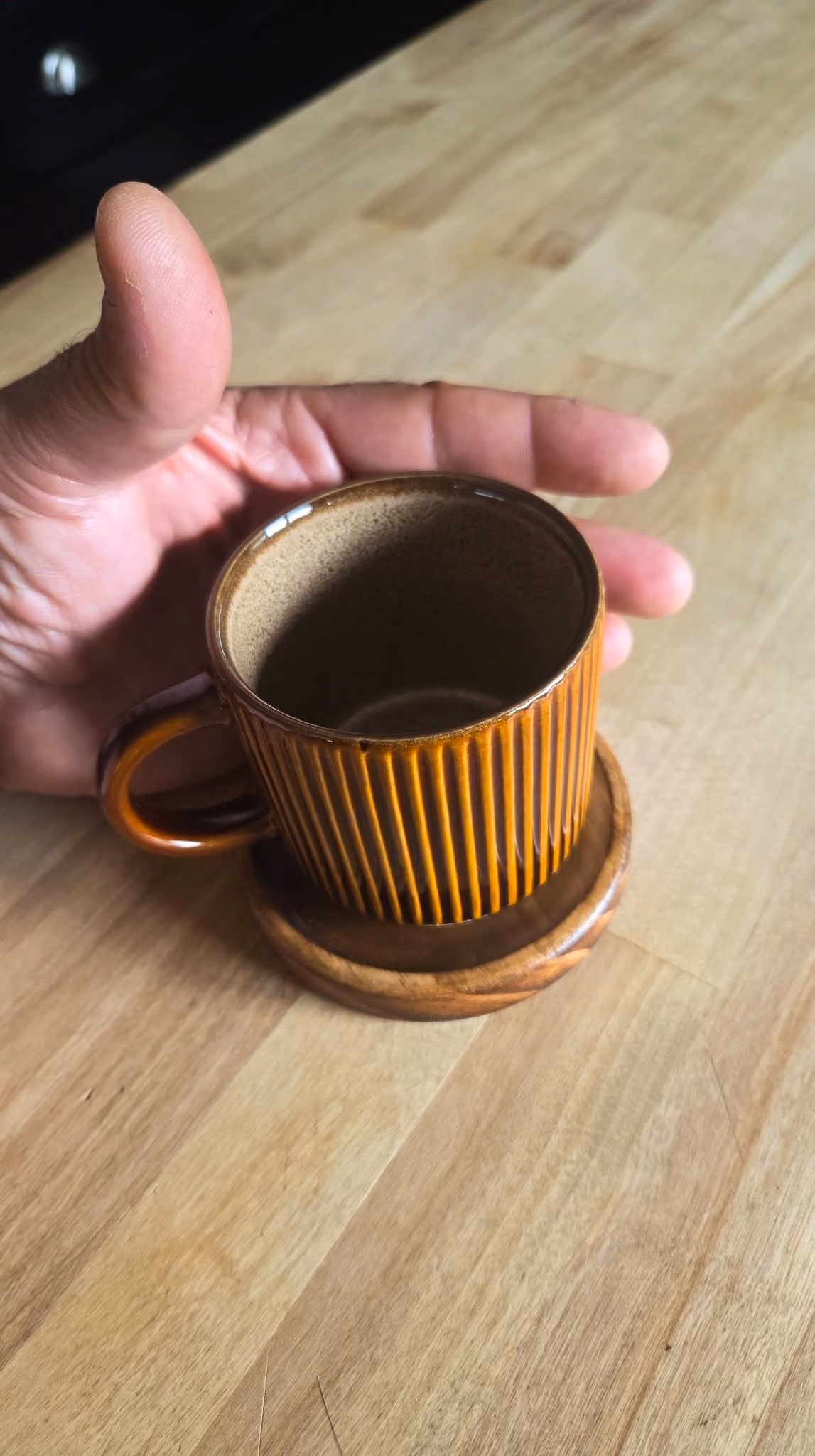 Hand holding a brown striped ceramic cup on a wooden saucer on a light wood table.