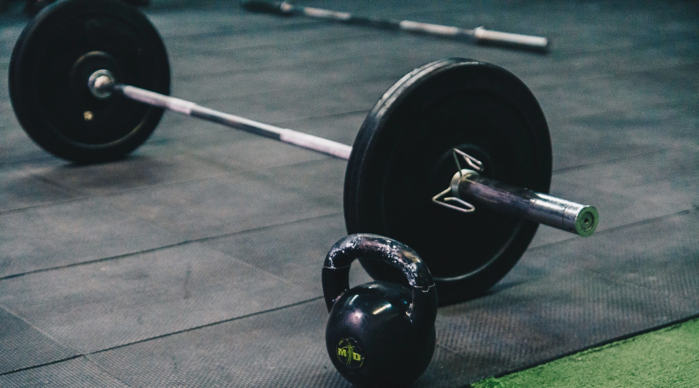 A loaded barbell with black bumper plates sits on a black rubber mat next to a worn black kettlebell, with another barbell resting in the background inside a gym.