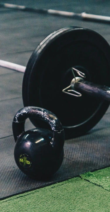 A loaded barbell with black bumper plates sits on a black rubber mat next to a worn black kettlebell, with another barbell resting in the background inside a gym.