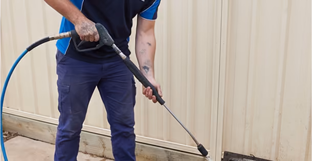 Person wearing blue pants and shirt holding a pressure washer spraying water on a wall.