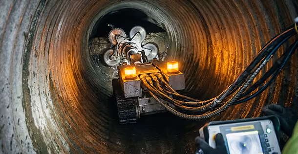 Robotic machine cleaning the inside of a large rusty metal pipe with a control device in view.