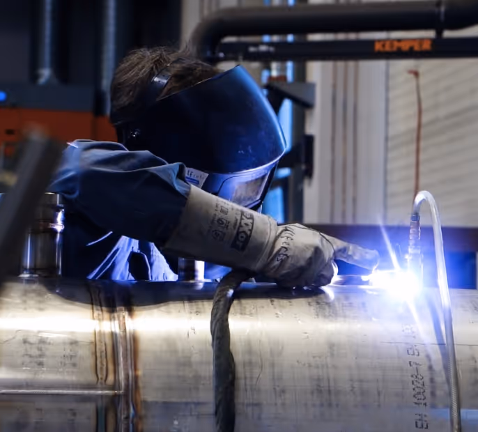 Ein Schweißer in Schutzhelm und Handschuhen arbeitet an einem Metallzylinder und erzeugt einen hellen Lichtfunken.