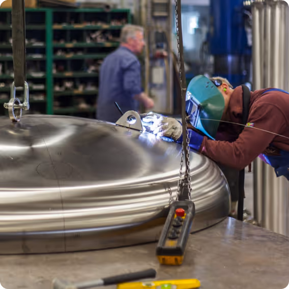 Ein Schweißer trägt einen Schutzhelm und arbeitet an einem großen, polierten Metallbehälter in einer Werkstatt.