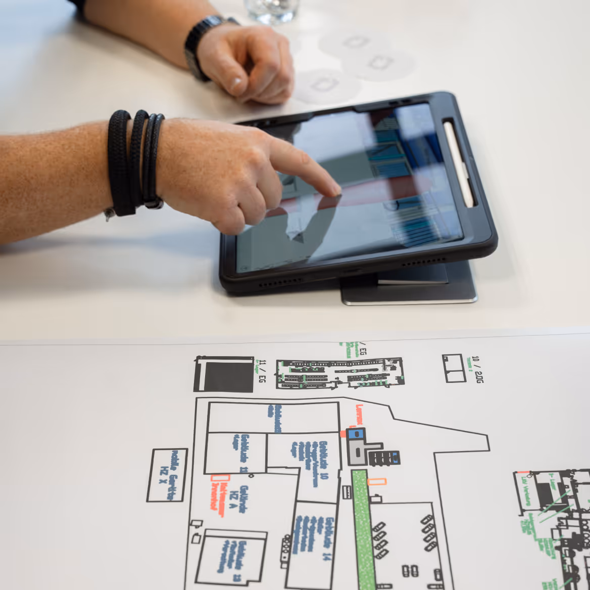 Hand zeigt auf ein Tablet mit einem Diagramm, das auf einem Tisch neben einem Bauplan liegt.