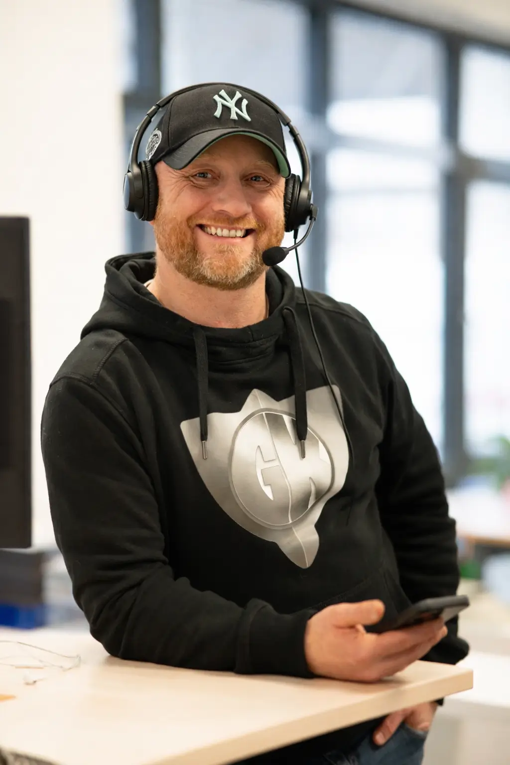 Mann mit schwarzer Kappe und Headset lächelt, trägt schwarzen Kapuzenpullover und hält ein Smartphone in der Hand.