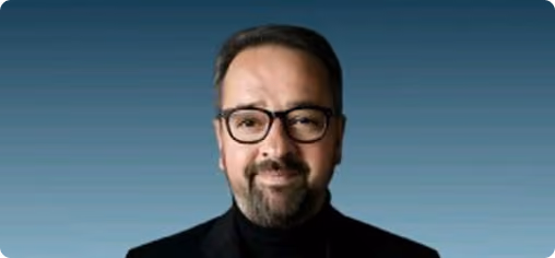 Portrait d'un homme souriant portant des lunettes noires et un col roulé noir sur fond bleu.