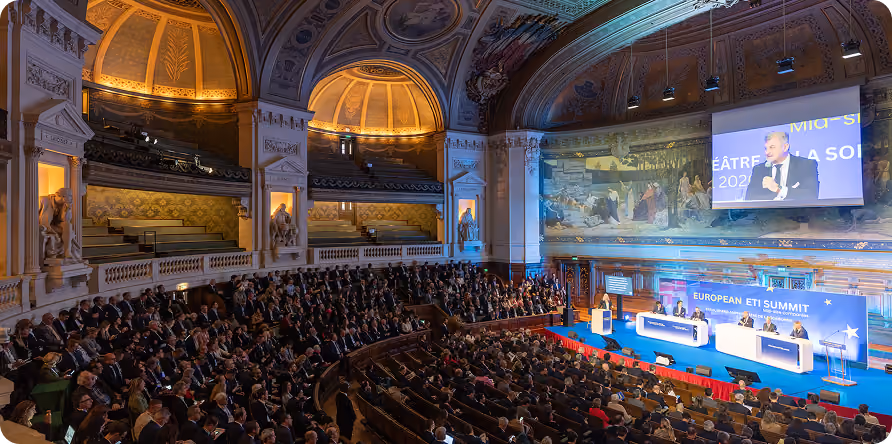 Conférence européenne dans un grand amphithéâtre ornée et pleine d'audience avec intervenants sur scène.