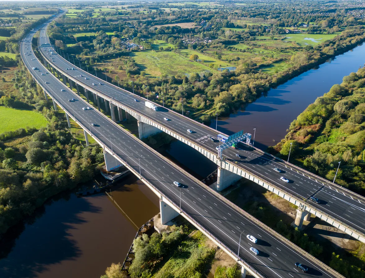 Aerial view of a large dual carriageway highway bridge crossing over a river surrounded by lush green fields and trees with scattered vehicles on the road.