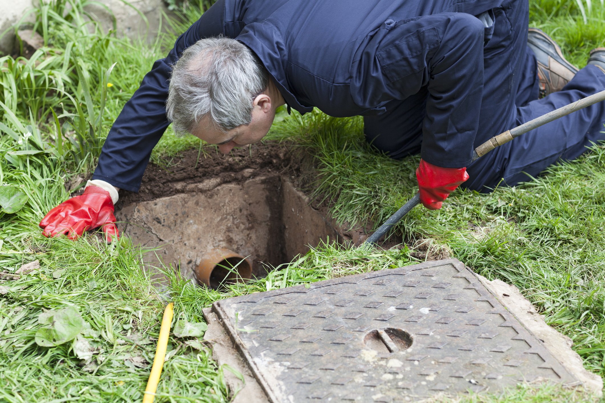Man wearing red gloves and dark blue work clothes kneeling on grass, inspecting a rectangular underground drainage or sewer hole with a visible pipe inside, holding a metal rod.