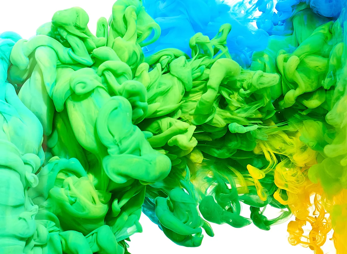 Swirling clouds of green, blue, and yellow ink mixing in water, creating abstract flowing shapes on a white background.