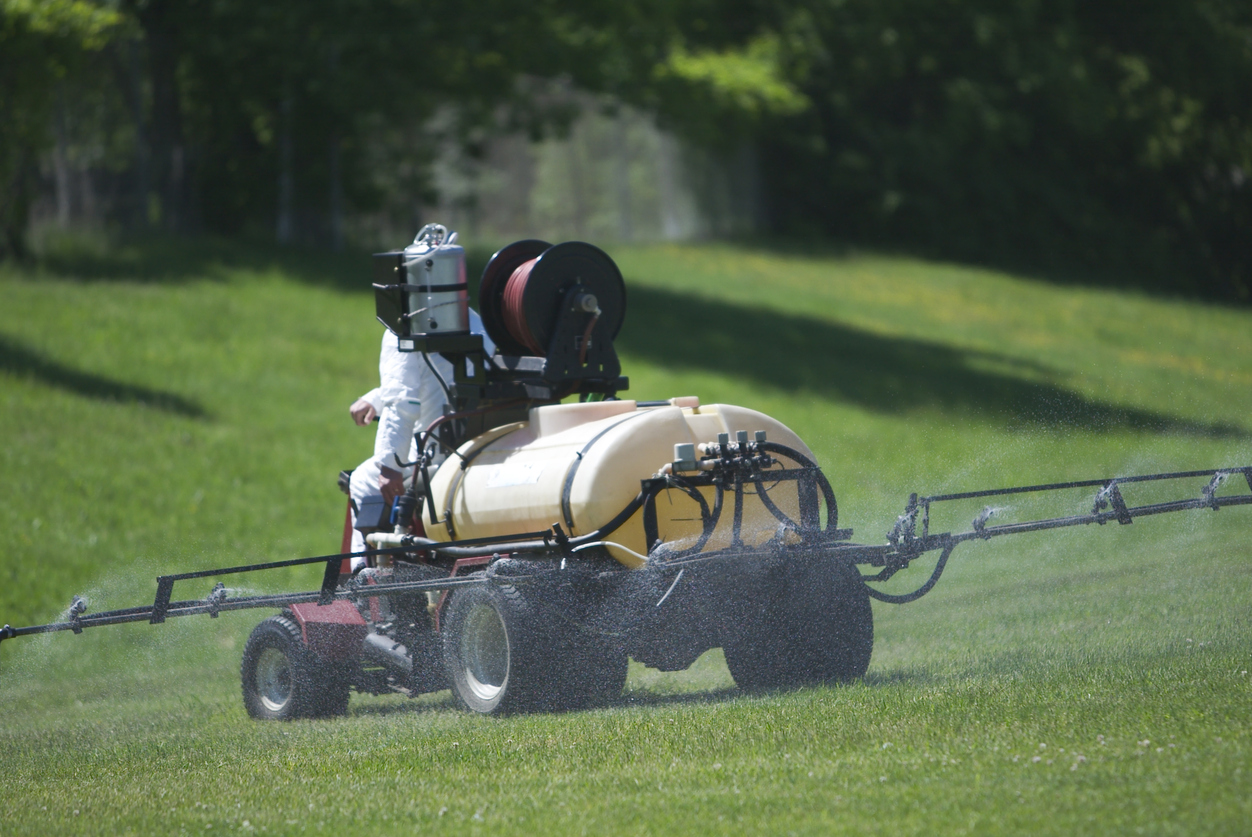 Person in protective white suit spraying a field with an agricultural sprayer mounted on a small tractor releasing mist over green grass under sunlight.