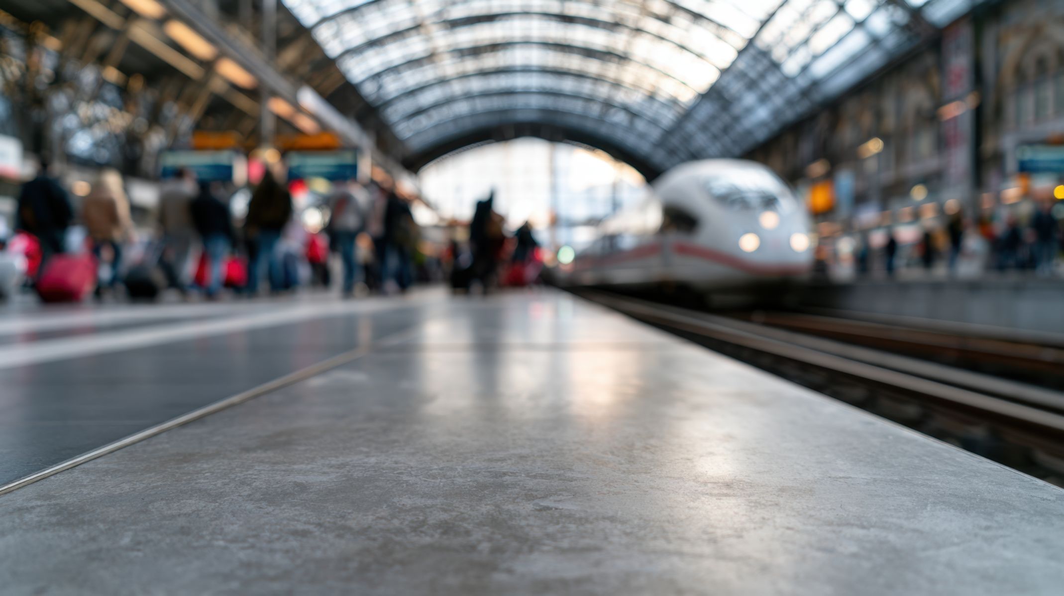 Blurred view of a busy train station platform with a modern white train on the tracks and passengers with luggage waiting under a glass-arched roof.