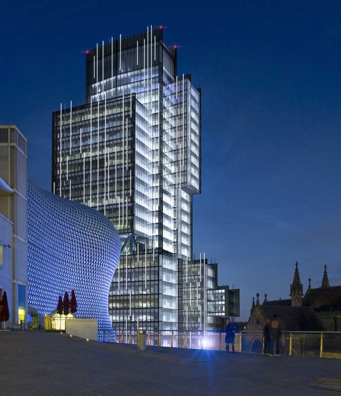 Modern glass skyscraper illuminated at night with vertical white light strips next to a building with a curved wall covered in circular metallic panels, and gothic architectural structures in the background under a clear dark blue sky.