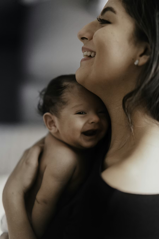 Smiling woman holding and cuddling a happy baby close to her neck indoors.