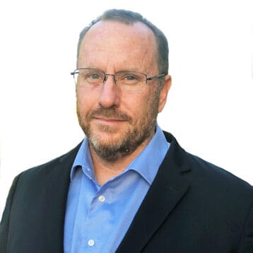 Man with glasses and beard wearing a blue shirt and black blazer on a white background.