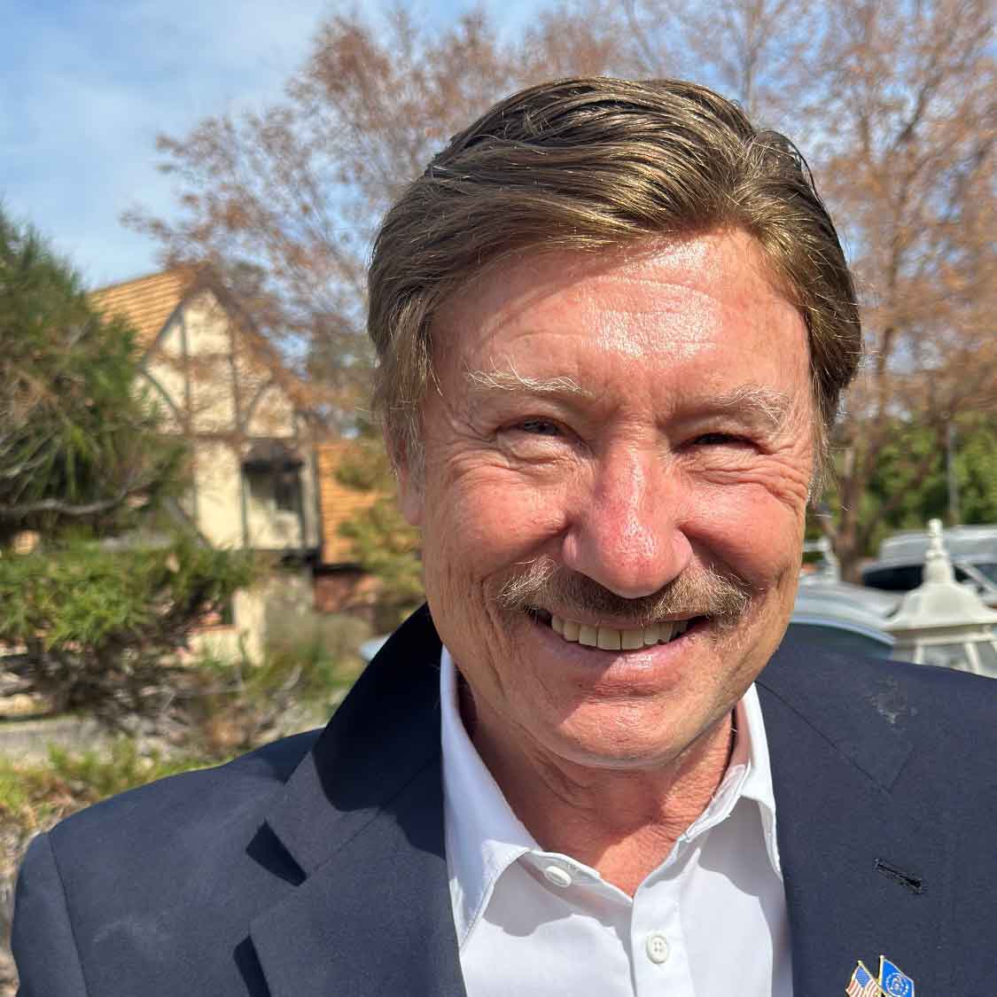 Smiling middle-aged man with mustache outdoors wearing a navy suit and white shirt.