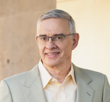 Smiling middle-aged man with glasses in beige suit jacket and light-colored shirt against neutral background.