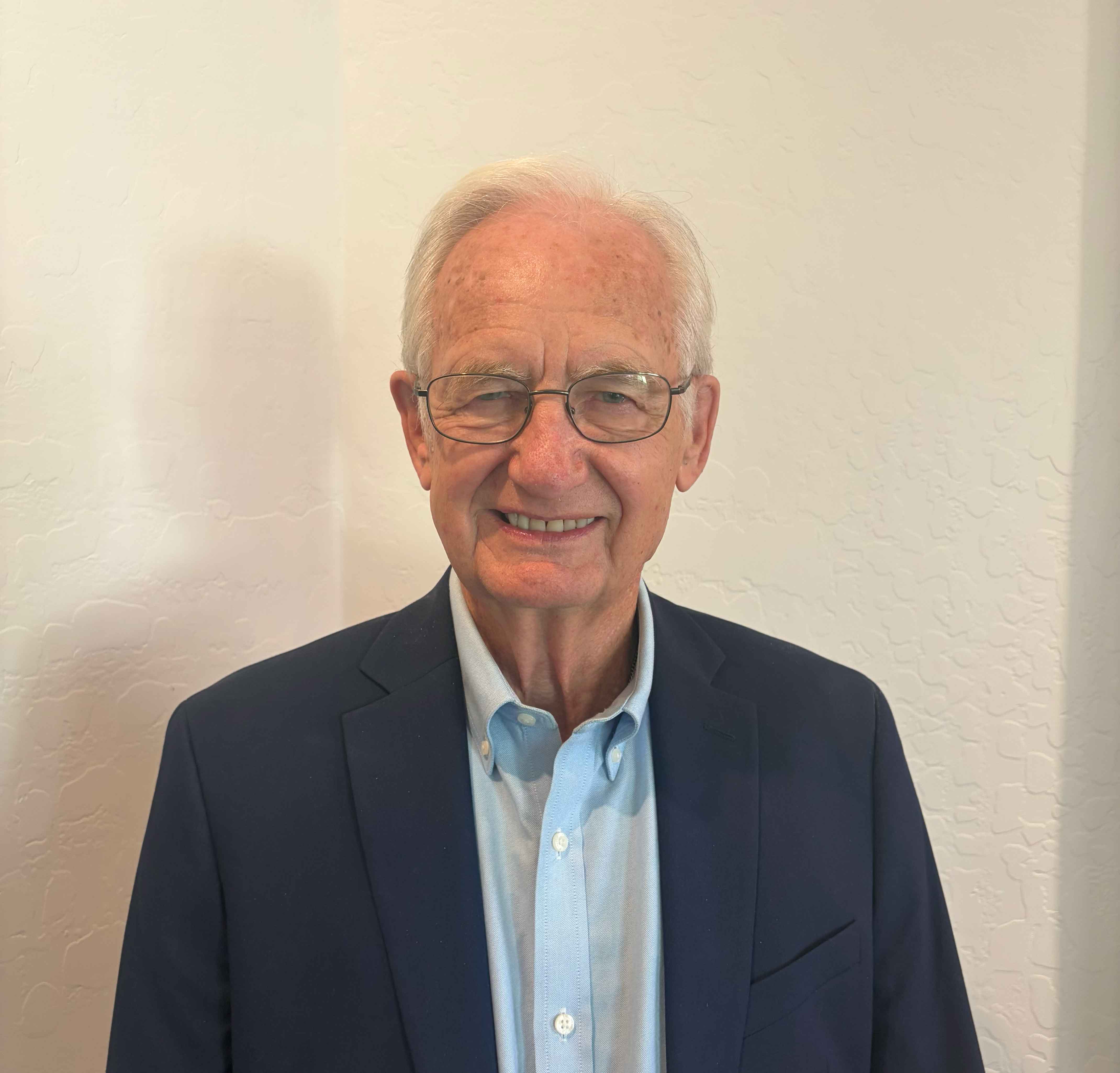 Smiling elderly man with glasses wearing a navy blazer and light blue shirt standing against a white wall.