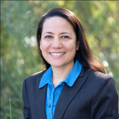Smiling woman with long dark hair in a blue collared shirt and dark blazer outdoors.