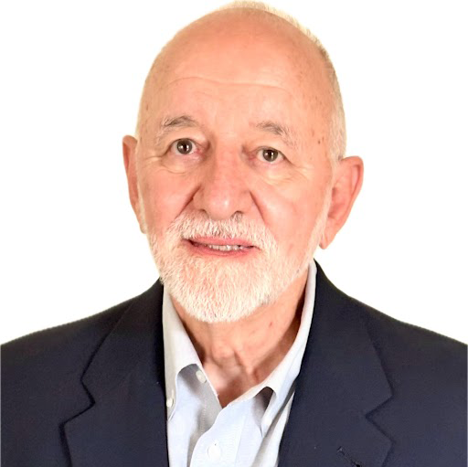 Older man with white beard and mustache wearing a dark blazer and light shirt against white background.