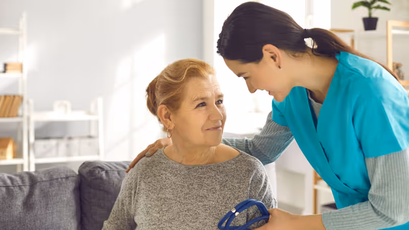 A healthcare worker in teal scrubs gently supports an elderly woman sitting on a gray sofa, offering a caring smile and holding a blue blood pressure cuff.