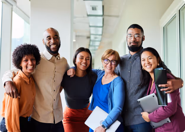 A diverse group of six smiling coworkers standing closely in an office corridor with large windows, some holding laptops and documents.