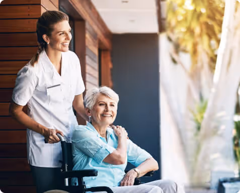 A smiling caregiver in white uniform stands behind an elderly woman in a wheelchair, both looking happy in a sunlit room with wooden walls and a large window with sheer curtains.