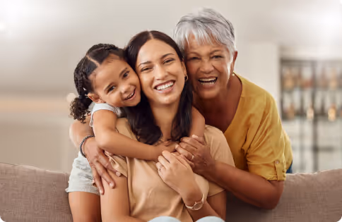 Three generations of women smiling and hugging on a sofa, including a young girl, a middle-aged woman, and an older woman with gray hair.