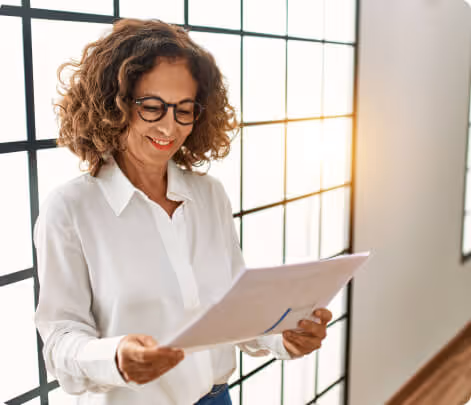Woman with curly hair and glasses wearing a white shirt, smiling while reading a document in a room with large grid windows.