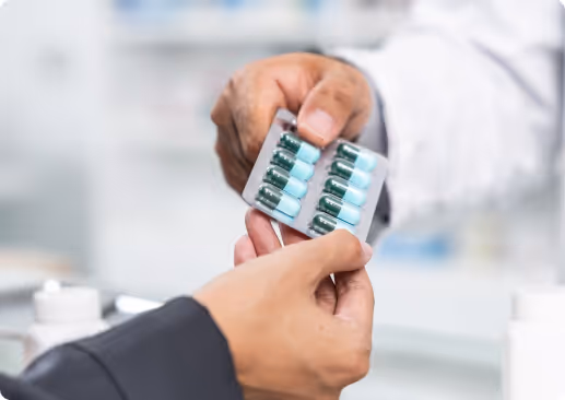 A pharmacist handing two blister packs of green and white capsules to a customer over the counter in a pharmacy setting.