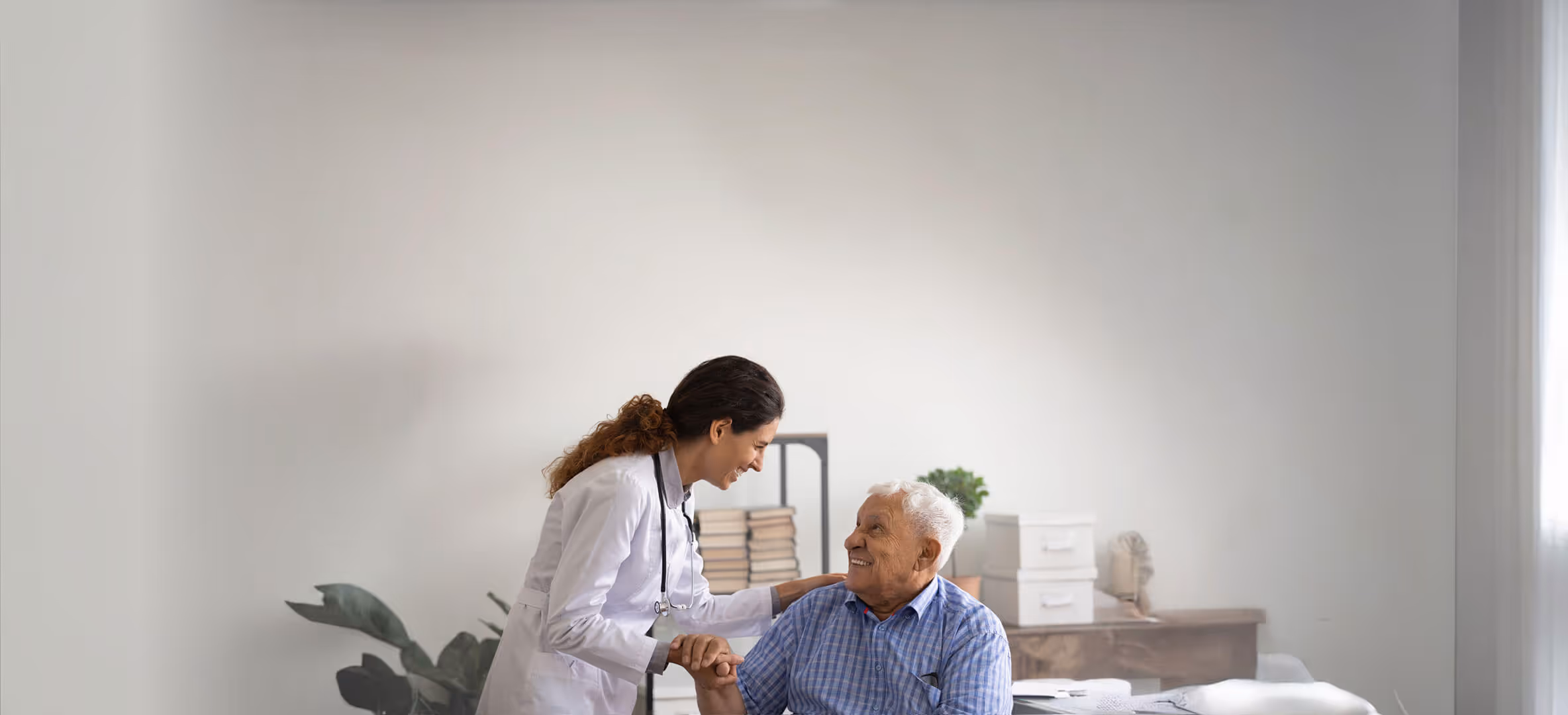 A female doctor in a white coat smiling and holding hands with an elderly man sitting on a bed in a bright, minimally decorated room.