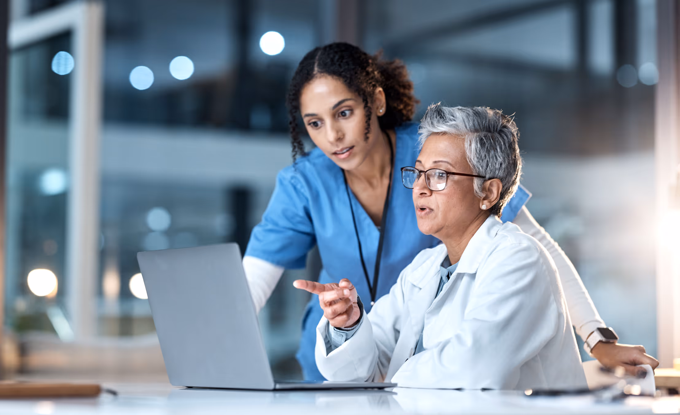 Two healthcare professionals, one older with gray hair and glasses in a white coat, and one younger in blue scrubs, looking at and discussing something on a laptop screen.