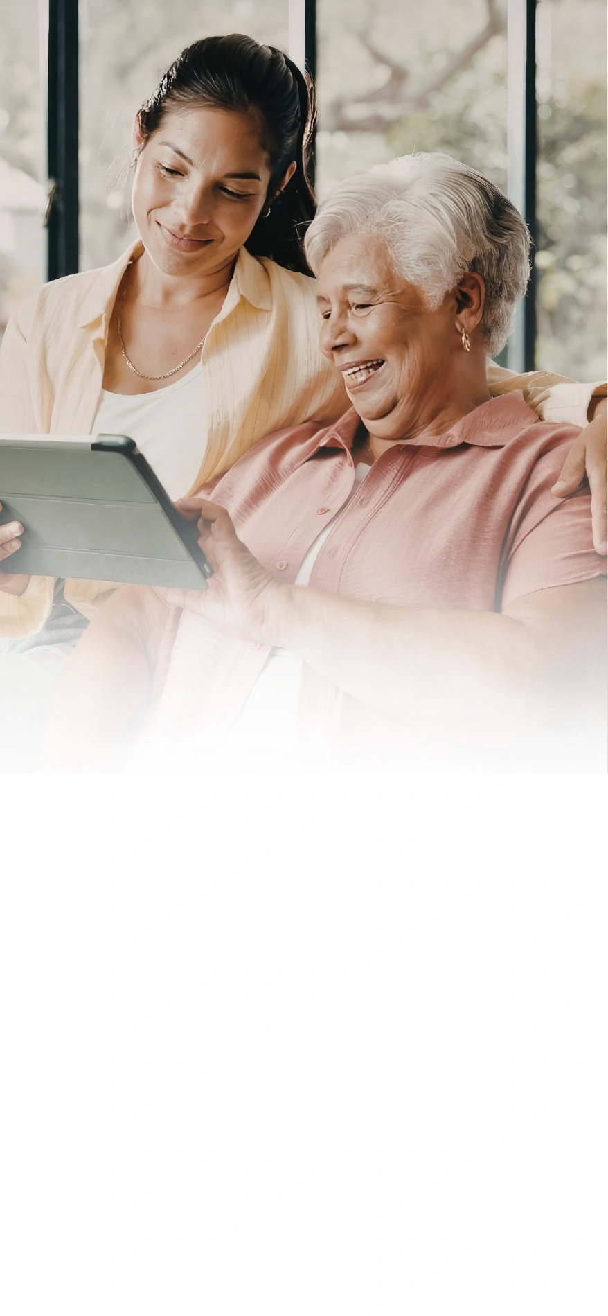 Younger woman and older woman sitting together on a couch, smiling and looking at a tablet the older woman is holding.