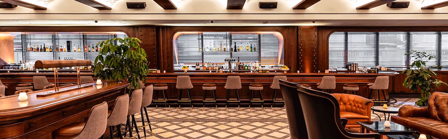 Luxury bar interior with wooden counters, upholstered stools, patterned floor, and leather seating areas.