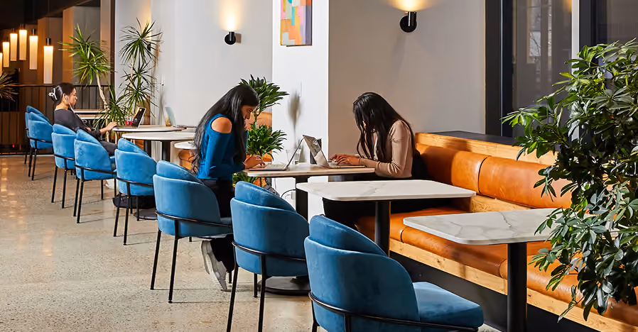 People working on laptops at tables with blue chairs and brown bench in a modern indoor space.
