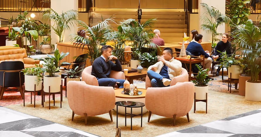 People sitting and conversing in a cozy indoor lounge with pink chairs and numerous potted plants.