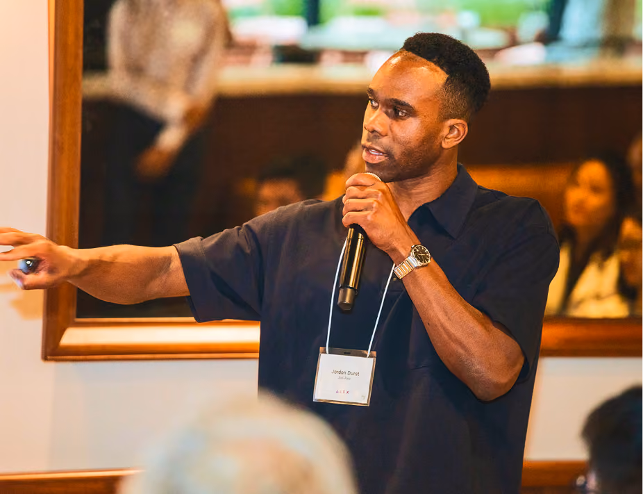 Man holding microphone and pointing, speaking to an audience indoors with a framed mirror behind him.