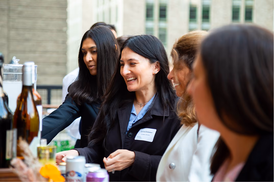Four women smiling and socializing around a table with drinks at an outdoor event.