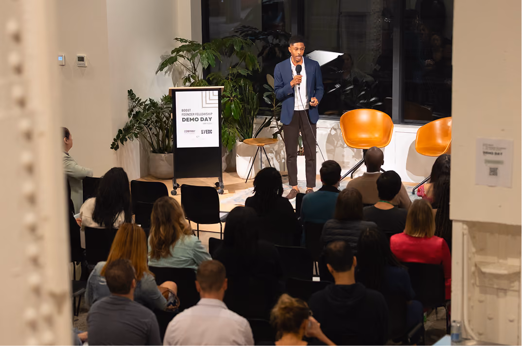 Man in blue jacket speaking into microphone at Boost Founder Fellowship Demo Day event audience facing him.