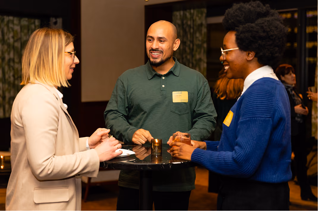 Three people standing and chatting around a small black table at a social event indoors.