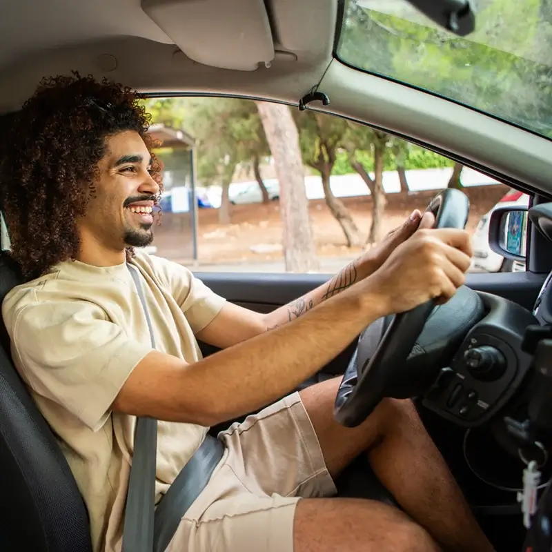 Man with curly hair smiling while driving a car wearing beige shorts and a t-shirt with seatbelt on.