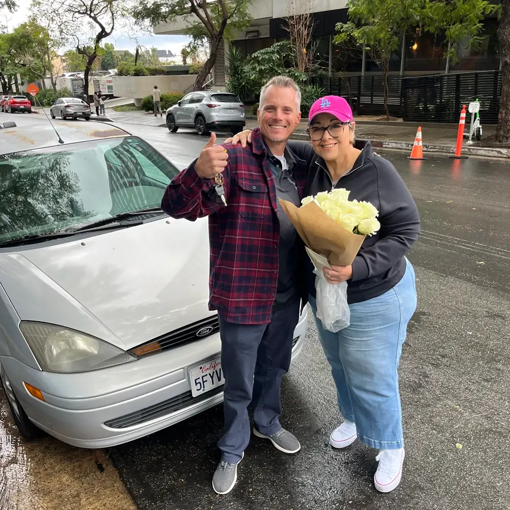 Jason Holland and Rae Ann standing beside the car she donated, holding the keys and a bouquet of flowers