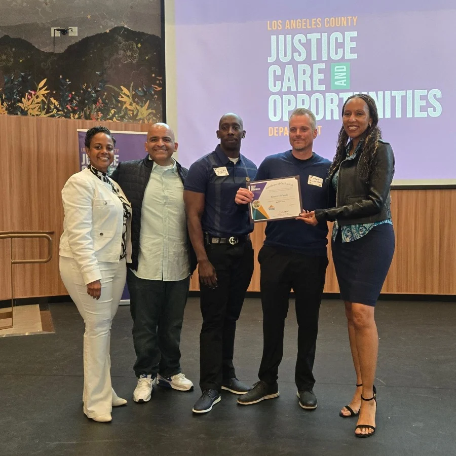 Five people standing on a stage holding a certificate at a Los Angeles County Justice, Care and Opportunities Department event
