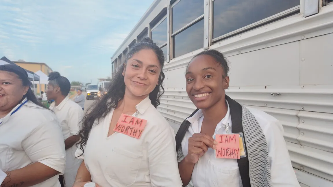 Two women holding 'I Am Worthy' signs by a bus