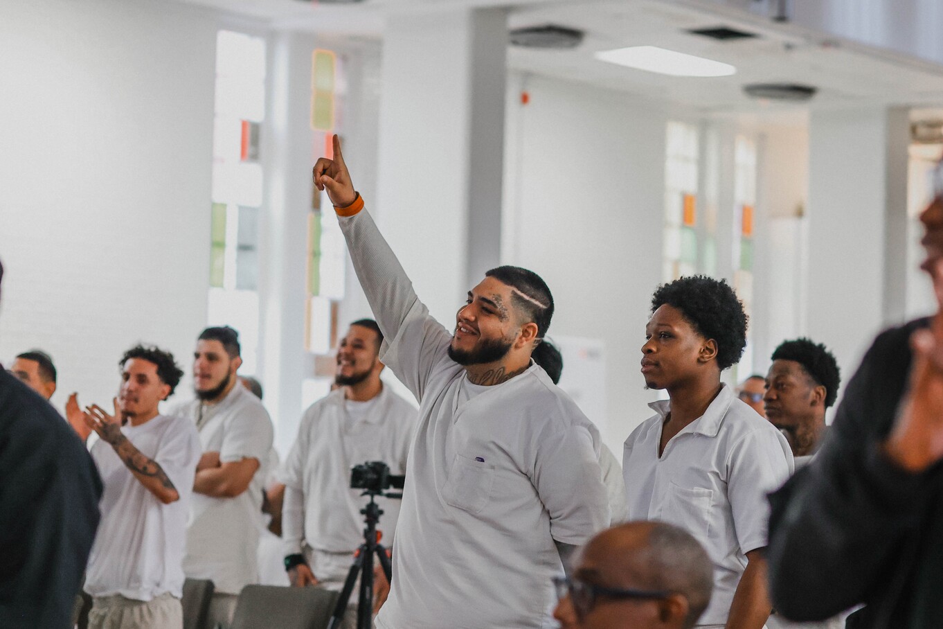 Man in white uniform raising hand and pointing during prison chapel service, surrounded by fellow inmates